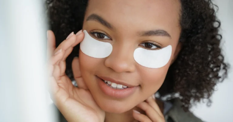 Smiling young woman with curly hair applying white under-eye patches for under-eye restoration in New York, NY, touching her face with both hands against a bright white background.